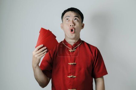 Cara conmocionada del hombre asiático que usa Cheongsam o tela tradicional china mientras se abanica a sí mismo usando angpao o regalo monetario rojo sobre fondo blanco. Gong Xi Fa Cai.