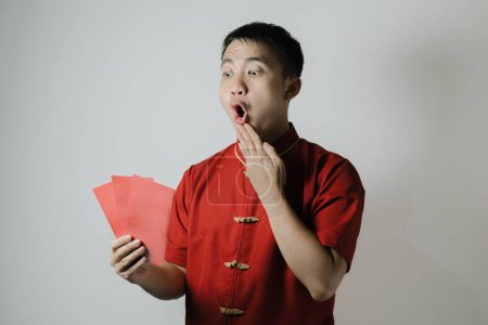 Téléchargez les photos : Visage choqué de l'homme asiatique portant Cheongsam ou tissu traditionnel chinois tout en tenant angpa ou cadeau monétaire rouge sur fond blanc. Gong Xi Fa Cai. - en image libre de droit