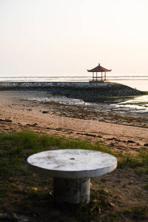 Beach scene at sunset with a traditional gazebo structure in the distance and an old round concrete table on the sandy shore