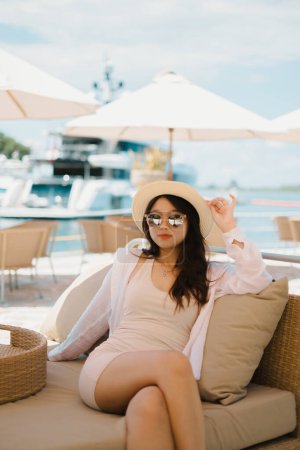 Photo for Beautiful young Asian girl is sitting in the cafe near the harbor while holding her straw hat with blurred yacht and sea in the background. Wearing slim cream dress, pink and reflective sunglasses, straw hat, and necklace. - Royalty Free Image