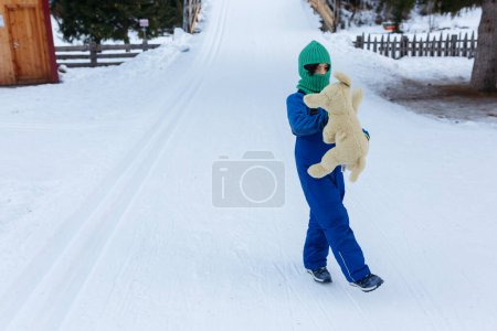 Photo for Child in skipants and green hat standing alone on snow holding her toy - Royalty Free Image