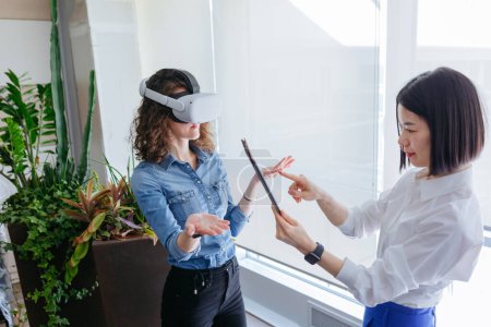 Photo for Two standing female colleagues in the office, one with curly hair wearing VR and another with short dark hair holding tablet - Royalty Free Image