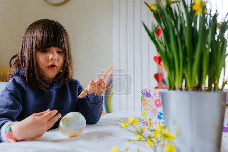 Foto de Sentado a la mesa divertido niño con capucha azul pintando el huevo en la cocina con el pincel - Imagen libre de derechos