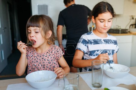 zwei kleine Schwestern essen zu Hause am weißen Tisch zu Mittag, während der Vater hinter ihr kocht