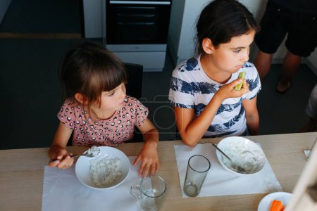 Ansicht von zwei kleinen Schwestern, die zu Hause am weißen Tisch zu Mittag essen, während der Vater im Hintergrund kocht