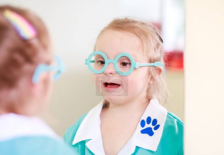 Photo for Portrait of a girl with Down syndrome. A girl in a doctor's playsuit. The child plays role-playing games. The girl looks in the mirror at her reflection. The baby is playing. - Royalty Free Image