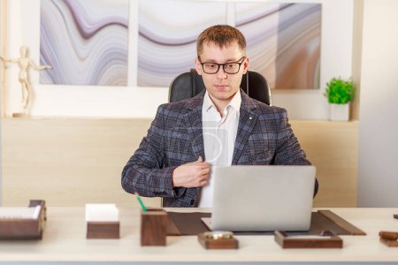 A businessman sits in the office at a large table in front of a laptop and works online, video chat, communication on working moments on the Internet, work from the office, the concept of remote work.