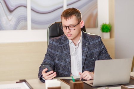 A businessman sits in the office at a large table in front of a laptop and works online, video chat, communication on working moments on the Internet, work from the office, the concept of remote work.