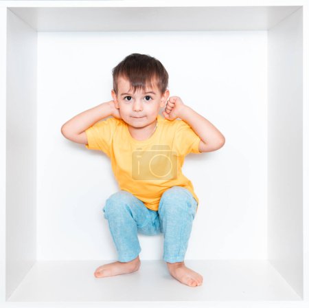 Un petit garçon mignon dans un T-shirt jaune et un pantalon bleu se trouve dans une niche blanche dans les meubles de sa chambre. Bébé dans une boîte carrée blanche.