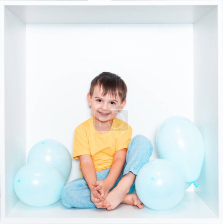 Un petit garçon mignon dans un T-shirt vert et pantalon bleu se trouve dans une niche blanche de meubles dans sa chambre. Bébé dans une boîte carrée blanche. Garçon avec des ballons bleus.