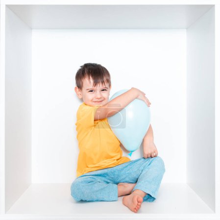 Un petit garçon mignon dans un T-shirt vert et pantalon bleu se trouve dans une niche blanche de meubles dans sa chambre. Bébé dans une boîte carrée blanche. Garçon avec des ballons bleus.