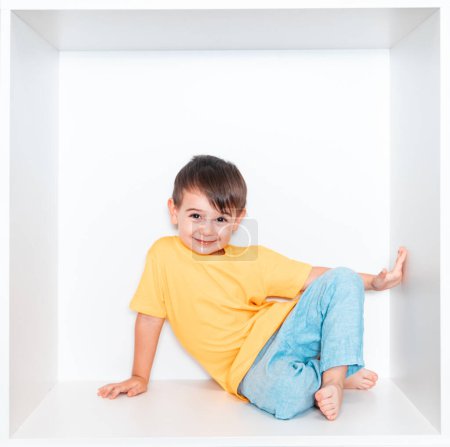 Un garçon mignon dans un T-shirt jaune et un pantalon bleu se trouve dans une niche blanche dans les meubles de sa chambre. Bébé dans une boîte carrée blanche.