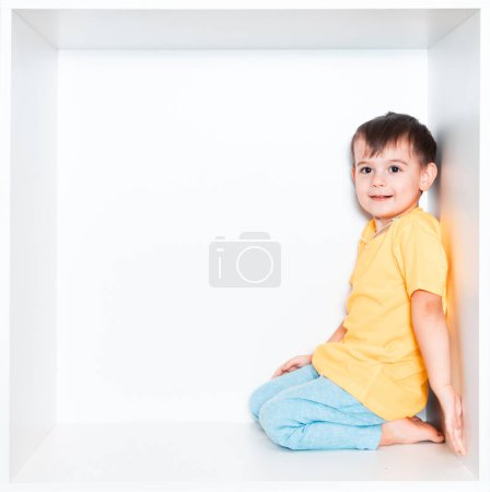 Un garçon mignon dans un T-shirt jaune et un pantalon bleu se trouve dans une niche blanche dans les meubles de sa chambre. Bébé dans une boîte carrée blanche.