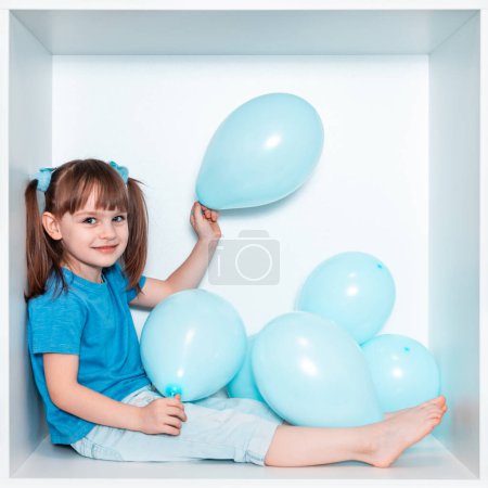 Une jolie petite fille en T-shirt bleu et pantalon bleu se trouve dans une niche blanche de meubles dans sa chambre. Bébé dans une boîte carrée blanche. Fille avec des ballons bleus.