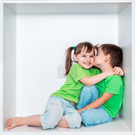 Une fille et un garçon en T-shirts verts jouent ensemble. Enfants sur fond blanc. Enfants dans une niche de meubles blancs. Photo émotionnelle, enfance heureuse, enfants - frère et s?ur.