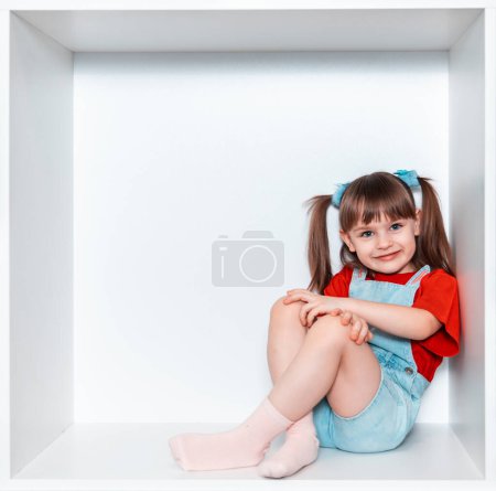 Une petite fille est assise dans le coin d'une grande boîte blanche en bois. Une fille en T-shirt rouge et short bleu. Magnifique enfance heureuse.