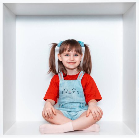  Une petite fille est assise les jambes croisées dans un cube de meubles blancs. L'enfant exprime des émotions joyeuses.