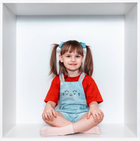 Une belle petite fille est assise dans une grande armoire blanche en forme de carré. La fille sourit très joliment, elle a un look expressif, le bébé porte un T-shirt rouge.