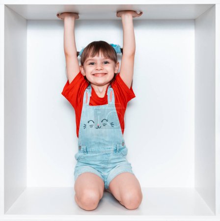 Une petite fille est assise dans une boîte sur ses genoux et lève les mains. Une fille en T-shirt rouge et short bleu. Beau sourire heureux sur le visage d'un enfant.