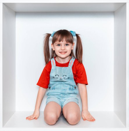 Une petite fille est assise dans une boîte sur ses genoux sur un fond blanc. Une fille en T-shirt rouge. L'enfant est monté dans l'armoire blanche. Bonne enfance.