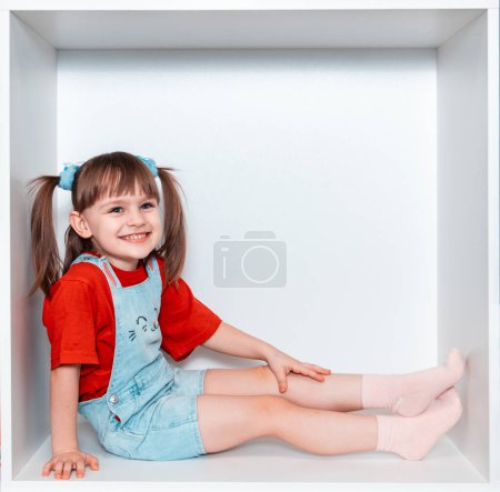Une petite fille est assise dans une boîte, sur un fond blanc. La fille a 3 ans. L'enfant est monté dans l'armoire blanche. Elle sourit magnifiquement. Bonne enfance.