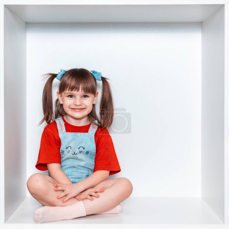 Une petite fille est assise dans une boîte, sur un fond blanc. La fille a 3 ans. L'enfant est monté dans l'armoire blanche. Elle sourit magnifiquement. Bonne enfance.