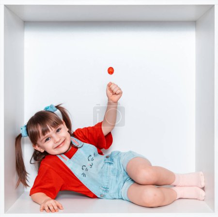  Une petite fille se couche sur le côté et tient une sucette rouge dans sa main. La fille a 3 ans. L'enfant sourit magnifiquement. Bonne enfance.