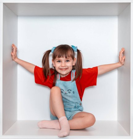 Une belle petite fille est assise dans une grande armoire blanche en forme de carré. La fille sourit très joliment, elle a un look expressif, le bébé porte un T-shirt rouge.
