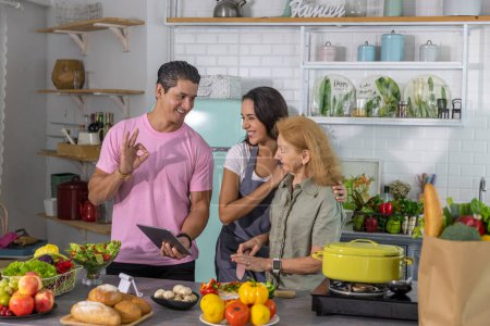 Photo for Young couple working with grand mom to take online cooking course and prepare meal at home. Happy family is cooking breakfast with healthy food together - Royalty Free Image