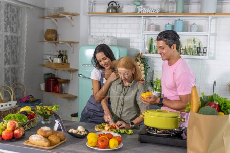 Photo for Young couple working with grand mom to take online cooking course and prepare meal at home. Happy family is cooking breakfast with healthy food together - Royalty Free Image
