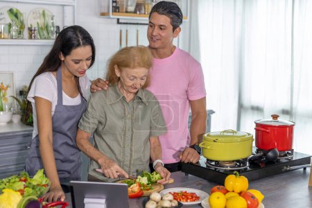 Photo for Young couple working with grand mom to take online cooking course and prepare meal at home. Happy family is cooking breakfast with healthy organic food together - Royalty Free Image