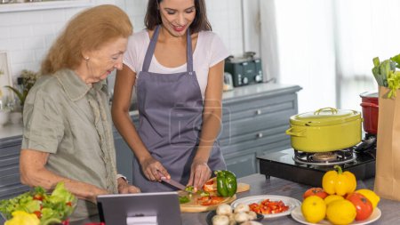 Foto de La madre anciana y su hija pequeña reciben clases de cocina en línea para preparar la comida en casa. Buena relación familiar entre mamá e hija cocinando juntas - Imagen libre de derechos