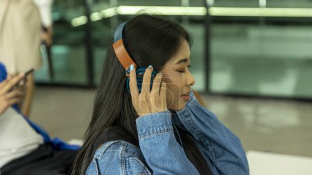 Foto de A la joven le gusta escuchar música con auriculares inalámbricos mientras espera en un salón del aeropuerto antes de abordar. Mujer viajera se siente feliz con la música esperando su viaje en una terminal de tren - Imagen libre de derechos