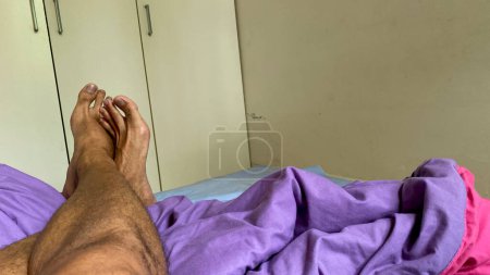 Photo for Asian man's leg. Foot Crossover relaxing on bed with white wardrobe on the background - Royalty Free Image