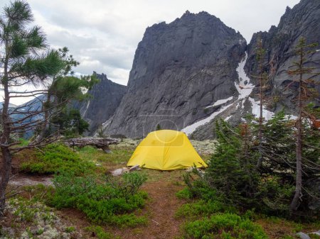 Dramatische Landschaft mit gelbem Zelt auf einem Waldhügel zwischen Felsen und sommerlicher Flora mit Blick auf große Felsen unter bewölktem Himmel. Einsames Zelt und grüne Sommerfarben im Hochgebirge.