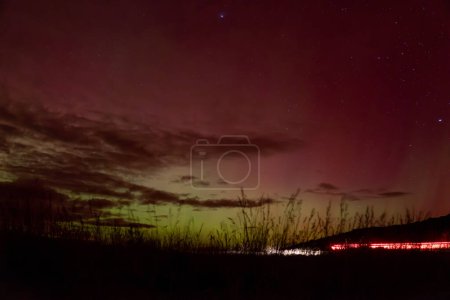 Ciel rouge et jaune de Aurora Australis avec des poutres roses au-dessus de Matakaea (Shag Point) dans l'île du Sud de la Nouvelle-Zélande. Un sentier lumineux des feux de voiture indique les nombreux spectateurs de cette aurore 2024.
