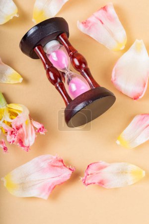 Withering Flower Petals Surrounding Hourglass on Beige Background, Top View