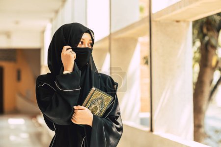 Junge junge muslimische Niqab-Frau, die im Gebäude der Universität den Koran und den Heiligen Al-Koran liest. Arabische schwarze Tschador-Dame.