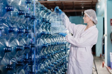 Junge kaukasische Arbeiterinnen arbeiten in der Trinkwasserfabrik und zählen den Vorrat an checkenden Wasserflaschen in Hygieneuniform am Arbeitsplatz