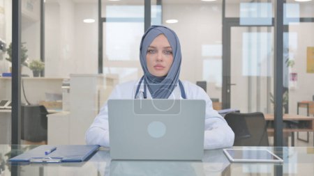 Arzt im Hijab blickt auf Kamera, während er am Laptop arbeitet