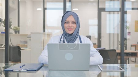 Médico en Hijab sacudiendo la cabeza en la aprobación mientras trabaja en el ordenador portátil