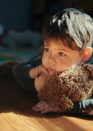 Nachdenklicher kleiner Junge umarmt zur Mittagszeit einen Spielzeugbär im Kinderzimmer