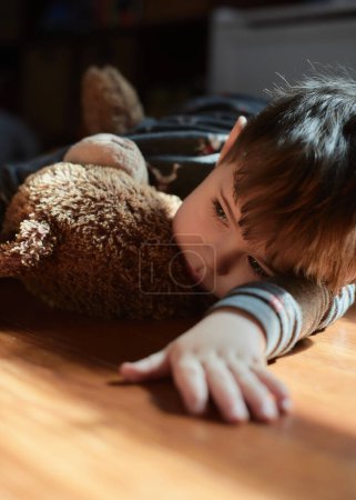 Nachdenklicher kleiner Junge umarmt zur Mittagszeit einen Spielzeugbär im Kinderzimmer
