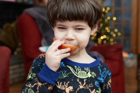 Foto de Un joven expresivo se está comiendo un tomate frente a un árbol de Navidad en la sala de estar - Imagen libre de derechos