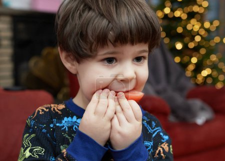 Foto de Un joven expresivo se está comiendo un tomate frente a un árbol de Navidad en la sala de estar - Imagen libre de derechos