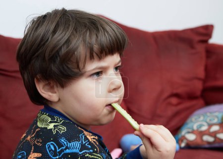 Foto de Un niño lindo bebé comiendo un pepino en el sofá en su pijama de dinosaurio - Imagen libre de derechos