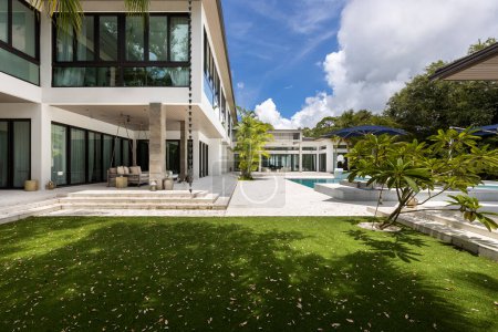 Elegante patio moderno con piscina y spa, sombrillas azules, plantas tropicales, palmeras, ventanas, patio cubierto con ventiladores de techo en el fondo y cielo azul