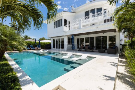 patio of elegant and modern house with swimming pool, surfboards, lounge chairs with umbrellas, palms, canal of miami bay, pier, boats,