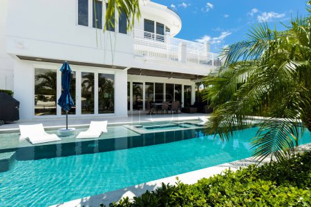 patio of elegant and modern house with swimming pool, surfboards, lounge chairs with umbrellas, palms, canal of miami bay, pier, boats,