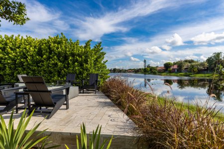 Lounge area overlooking the canal in Pembroke Pines, Fire pit with outdoor lounge chairs facing the water, short grass, blue sky, tropical climate, privet wall